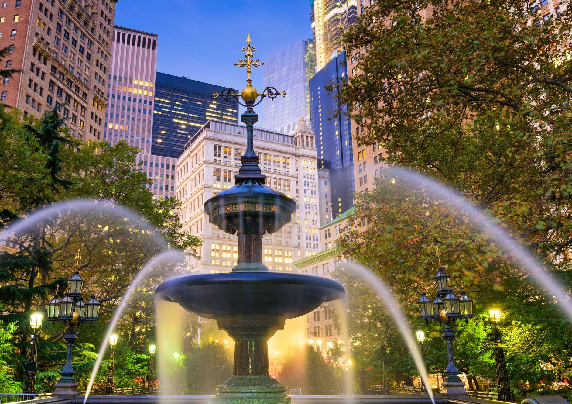 A large fountain in a park with a city skyline in the background.
